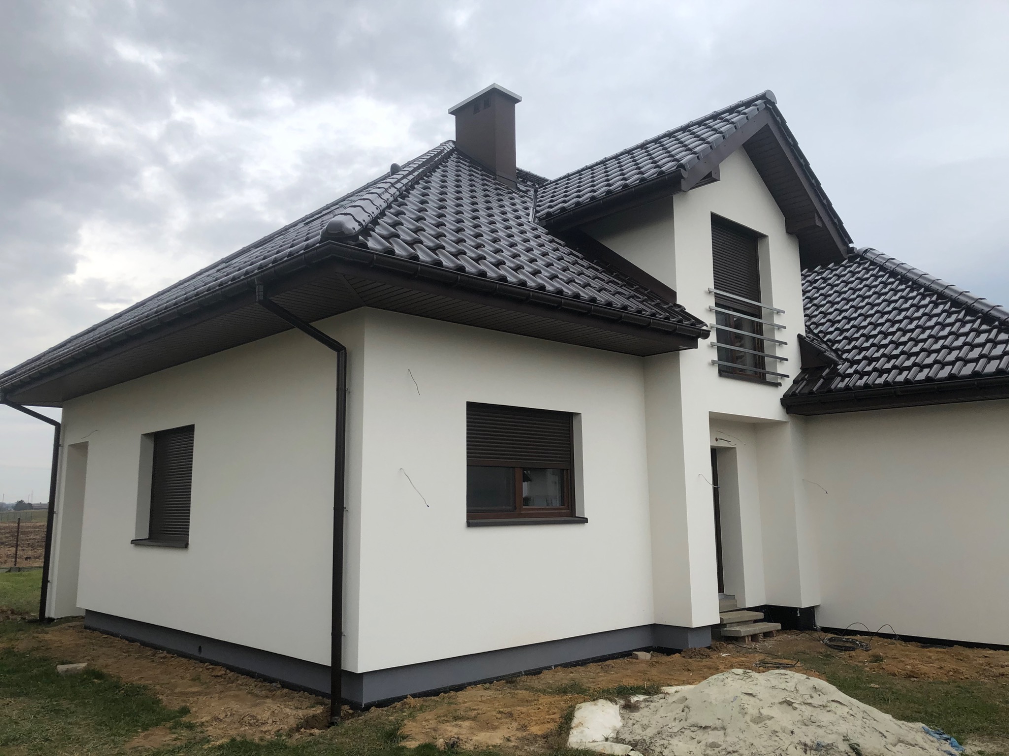 Biały dom jednorodzinny z brązową dachówką, szarym podmurowaniem i częściowo opuszczonymi roletami zewnętrznymi, widoczny z boku pod pochmurnym niebem.