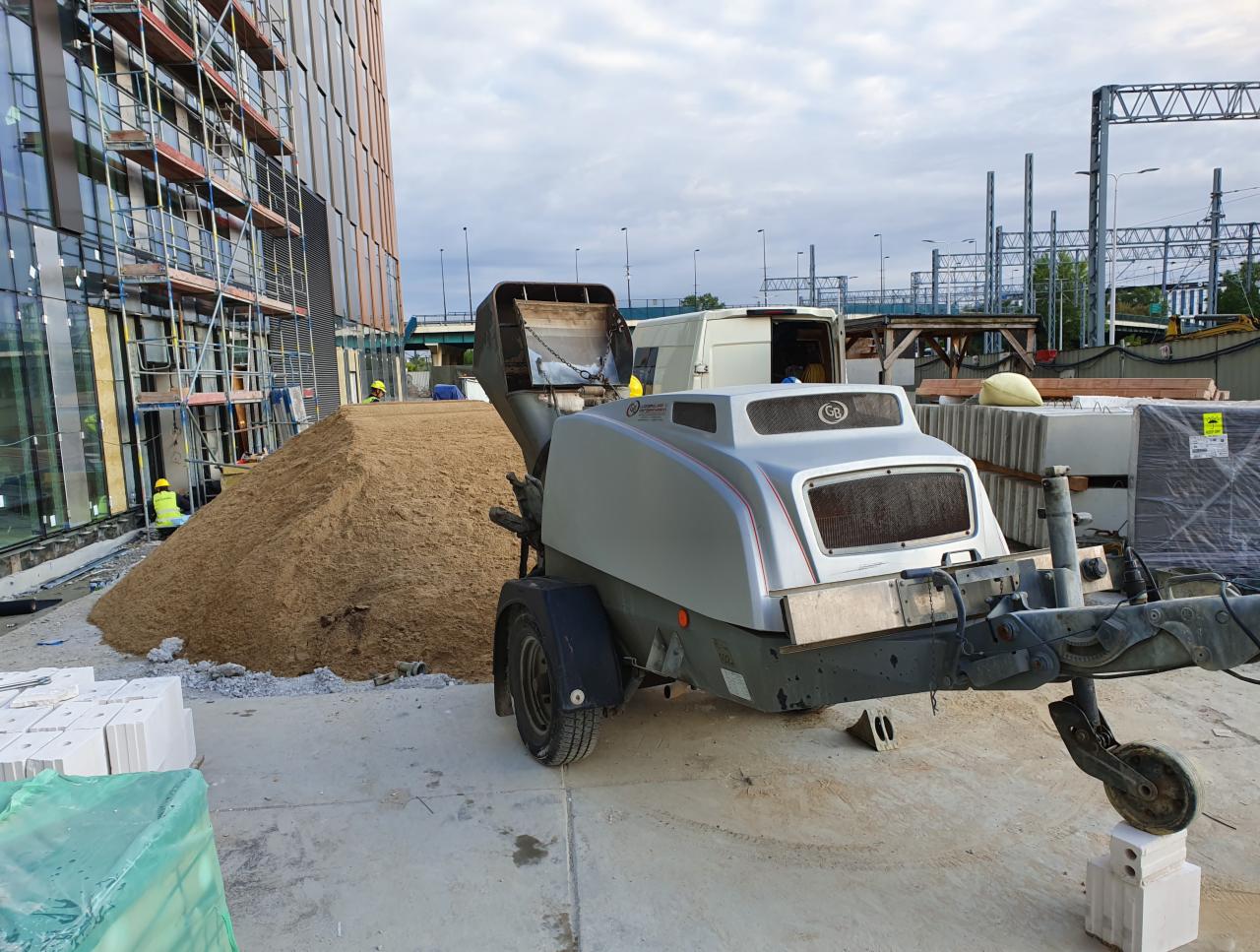 Agregat do wylewek stoi obok pryzmy piasku na budowie, w tle budynek w rusztowaniach i wiadukt kolejowy.