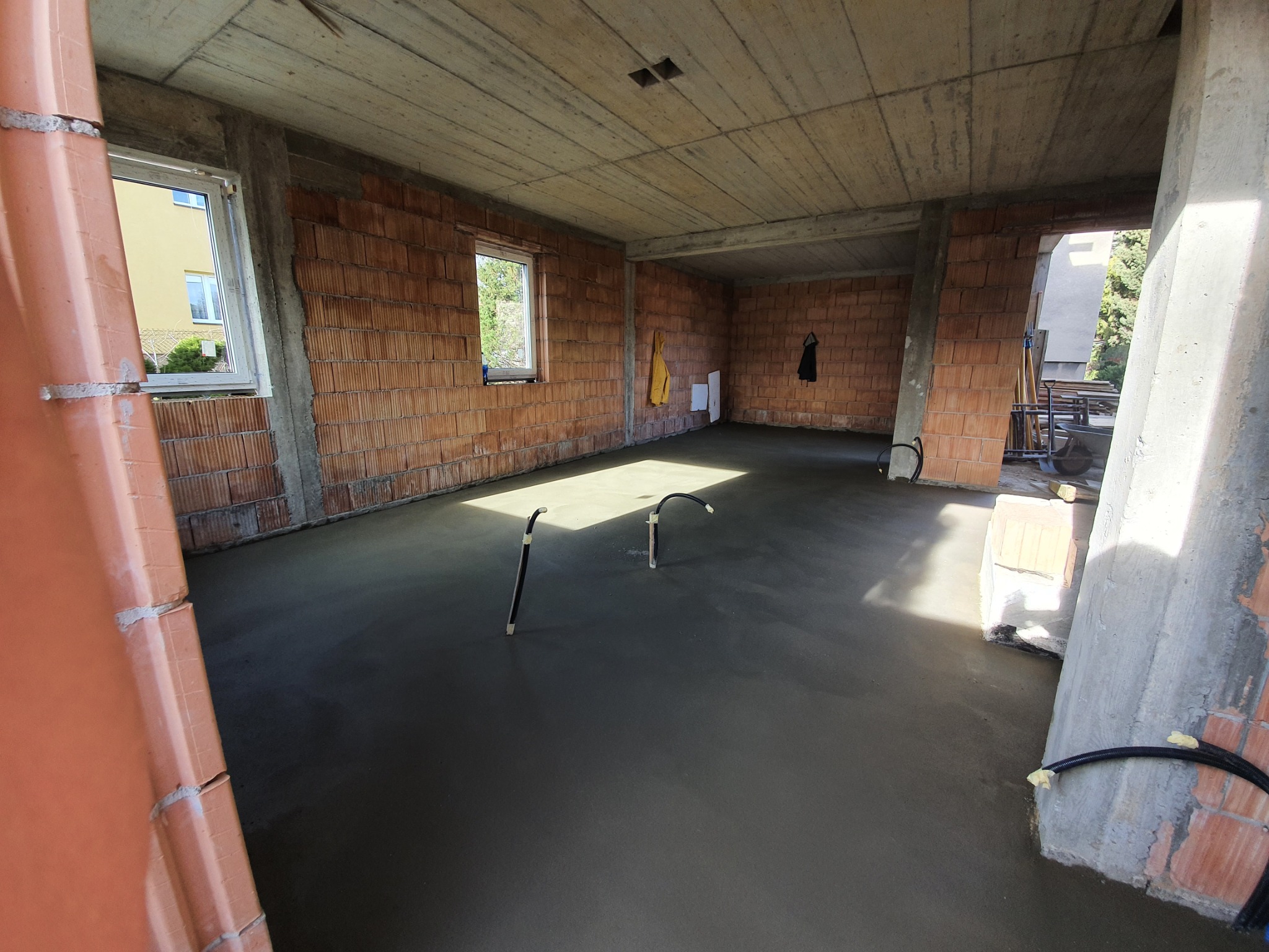 Świeżo wylany podbeton w surowym wnętrzu budynku z widocznymi przewodami instalacyjnymi wystającymi z posadzki, ściany z cegły kratówki i surowy betonowy strop.