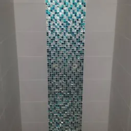 Pionowy pas mozaiki z turkusowo-srebrnych płytek w kabinie prysznicowej, z widocznymi dwoma otworami na baterię.