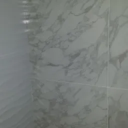 Fragment ściany w rogu pomieszczenia, wyłożonej płytkami imitującymi marmur oraz płytkami z delikatnym, falistym wzorem, widoczne ślady prac wykończeniowych na podłodze.