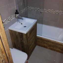 Nowoczesna łazienka z wanną i umywalką w szafce z ciemnego drewna, szare płytki z dekoracyjnym pasem mozaiki, widoczna część toalety po lewej stronie.