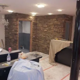 Ściana wyłożona dekoracyjnym kamieniem w odcieniach brązu i beżu, widoczne dwa otwory drzwiowe, w jednym z nich szara łazienka w trakcie prac, na podłodze folia ochronna, obok butelka wody i puszka...