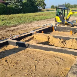 Przygotowanie terenu pod budowę domu: wykopane fundamenty z czarną izolacją, piasek wewnątrz, żółta mini koparka Yanmar i zagęszczarka w tle.