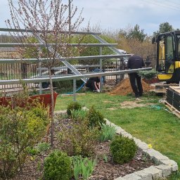 P.T.H.U. Multi-Bud - Montaż konstrukcji z metalowych profili w ogrodzie, widoczna koparka Yanmar, paleta z panelami, w tle dom i samochód