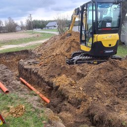 P.T.H.U. Multi-Bud - Żółta mini koparka Yanmar na usypanej ziemi obok wykopu z pomarańczowymi rurami kanalizacyjnymi, w tle domy i ogrodzenie.