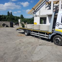 P.T.H.U. Multi-Bud - Laweta z napisem '24h POMOC DROGOWA' przewozi betonowe kręgi na tle budowy domu z białych bloczków.