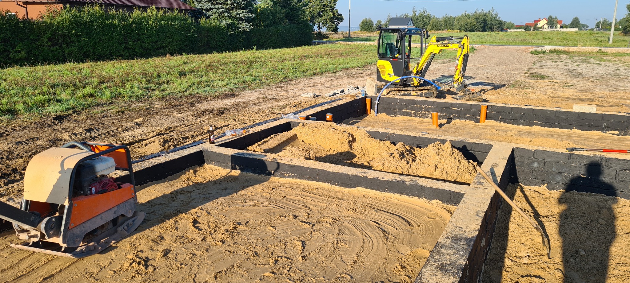 Przygotowanie terenu pod budowę domu: wykopane fundamenty z czarną izolacją, piasek wewnątrz, żółta mini koparka Yanmar i zagęszczarka w tle.