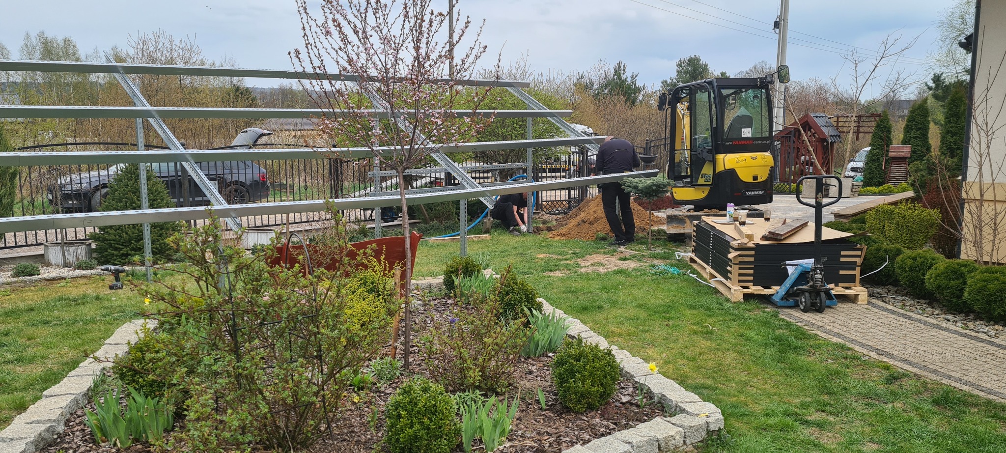 Montaż konstrukcji z metalowych profili w ogrodzie, widoczna koparka Yanmar, paleta z panelami, w tle dom i samochód