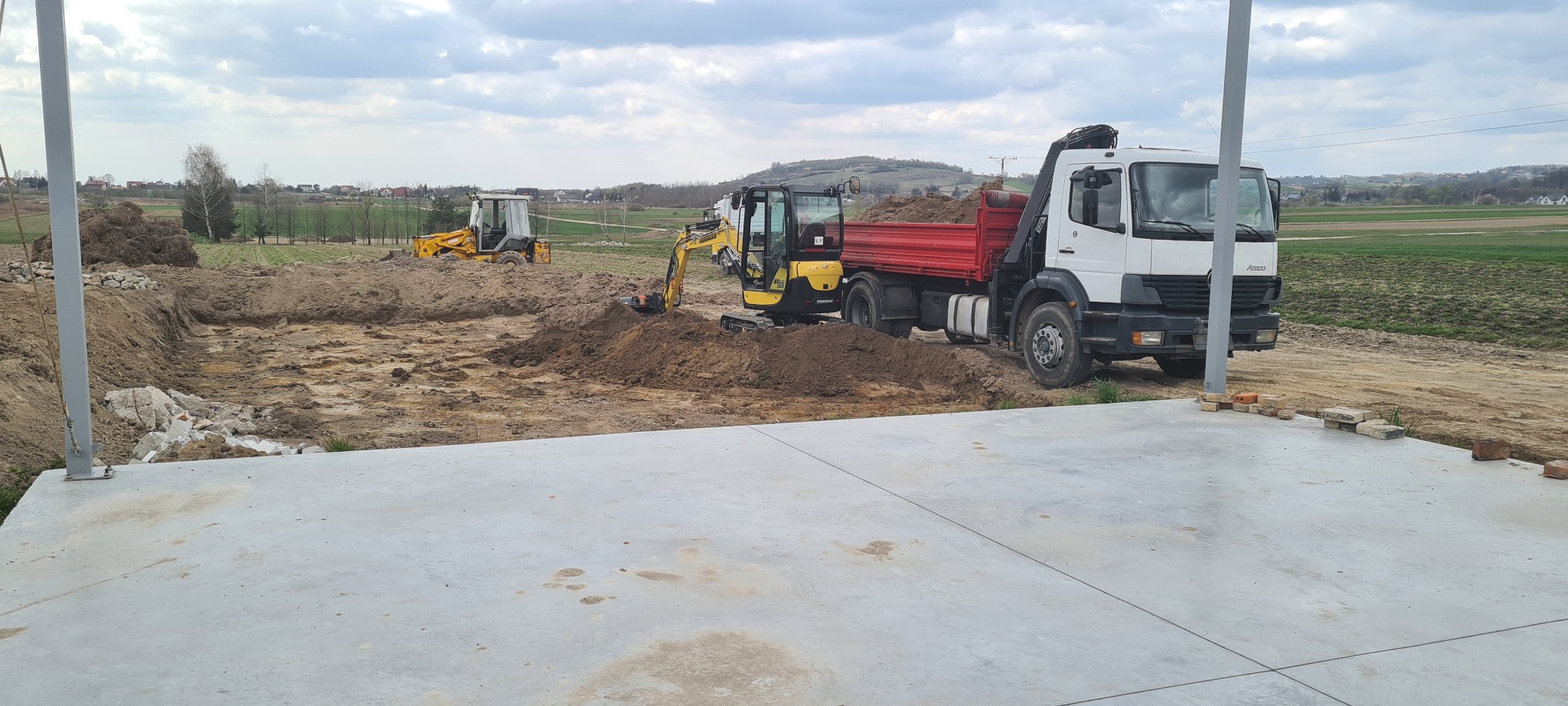Przygotowanie terenu pod budowę: wykop pod fundamenty, dwie koparki (żółta i czarna) oraz ciężarówka z czerwoną skrzynią ładunkową na tle zielonych pól i pochmurnego nieba.
