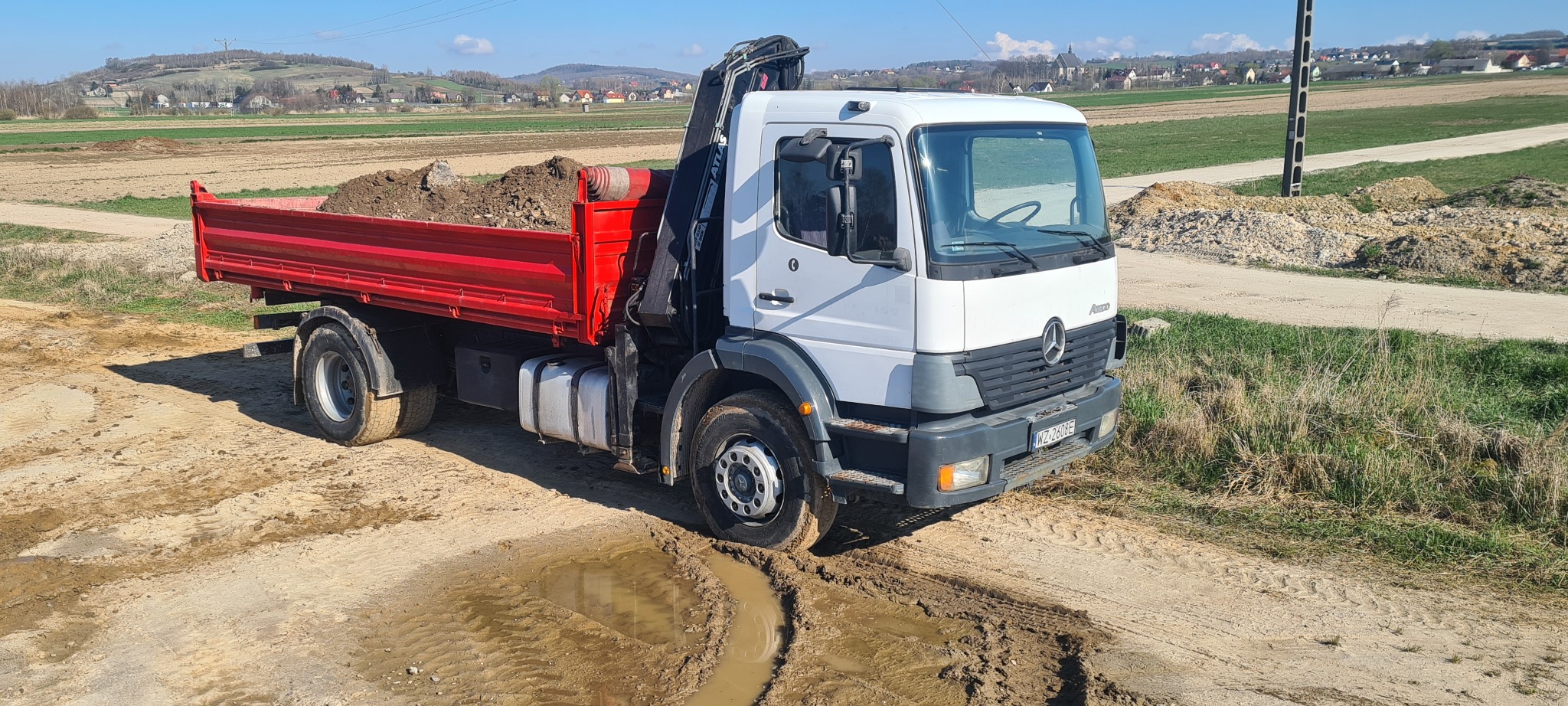 Ciężarówka Mercedes z czerwoną skrzynią załadowana ziemią stoi na błotnistym terenie budowy, w tle widoczne pola i zabudowania.