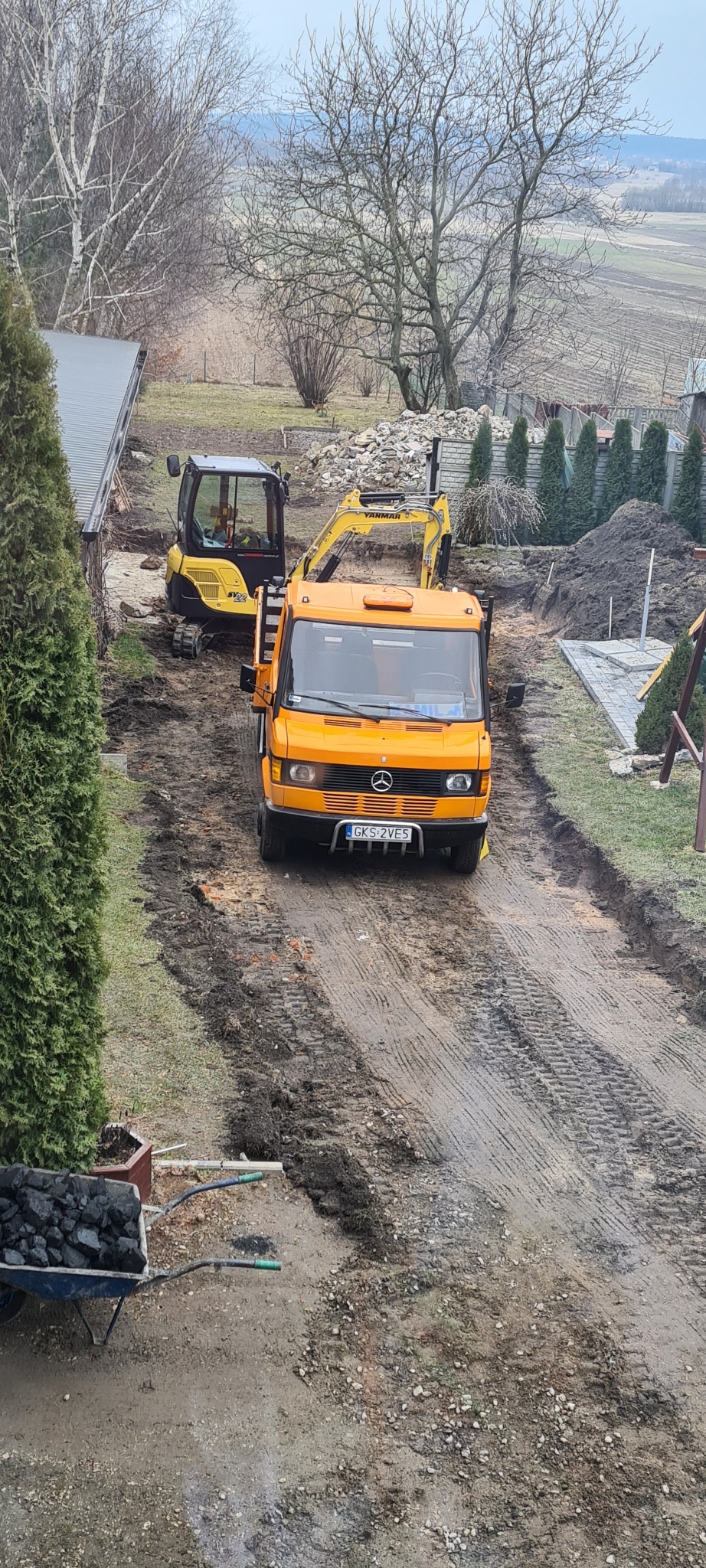 Pomarańczowy samochód dostawczy Mercedes parkuje na błotnistej drodze pomiędzy dwoma żółtymi minikoparkami; w tle widoczne wykopane rowy i hałdy ziemi, a także wózek z węglem kamiennym.