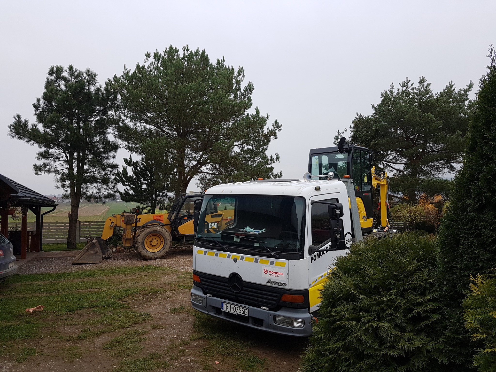 Żółta ładowarka CAT obok białej ciężarówki pomocy drogowej Mondial Atego na trawiastym terenie, z widocznymi drzewami i fragmentem budynku w tle.