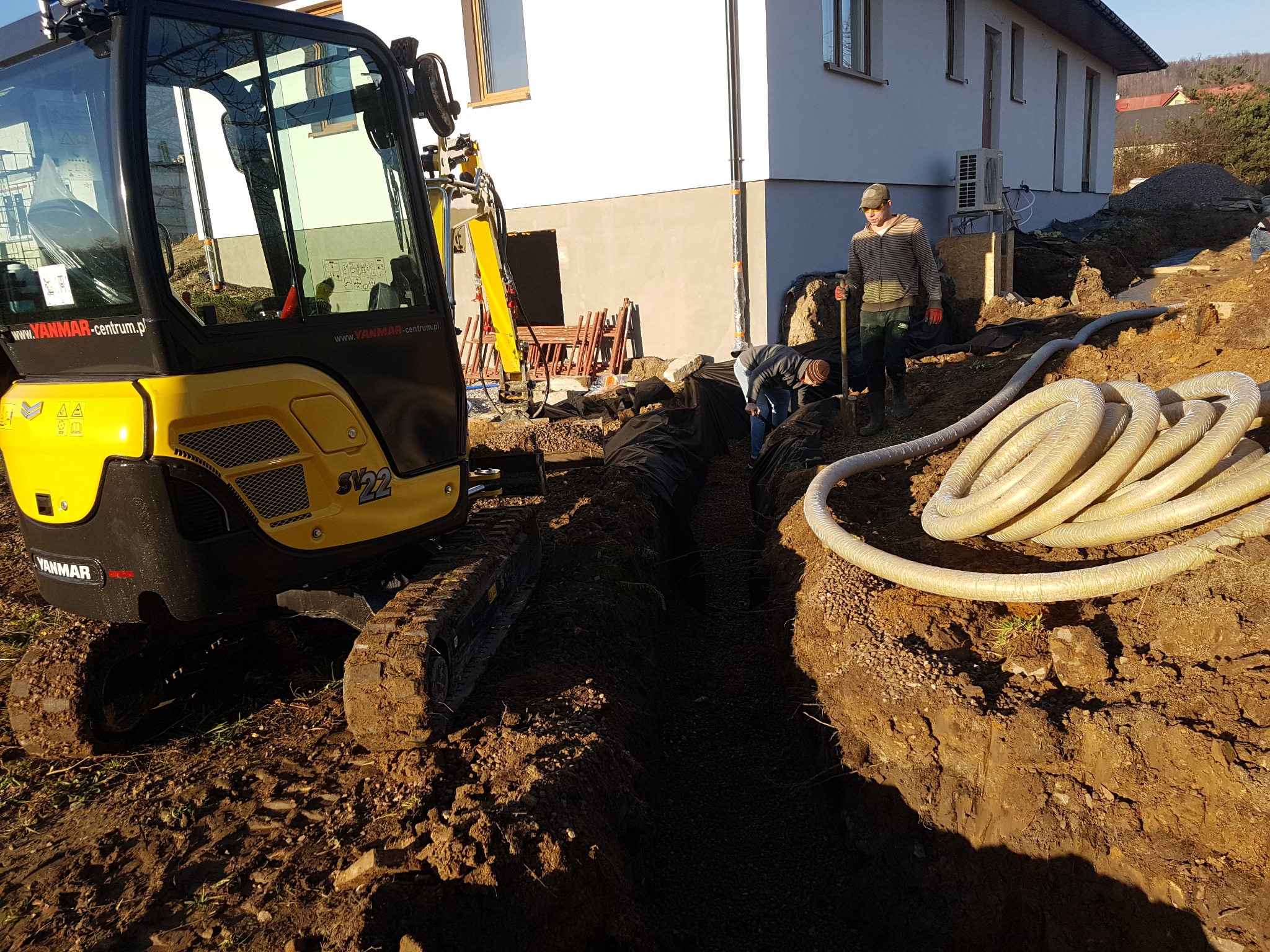 Żółta mini koparka Yanmar SV22 na placu budowy, obok wykopu z widoczną czarną folią, dwóch pracowników i zwiniętych rur wentylacyjnych, w tle budynek mieszkalny.