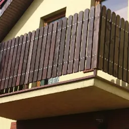 Drewniana balustrada balkonowa z pionowych desek, połączonych białymi nitami, na balkonie budynku z żółtą elewacją.