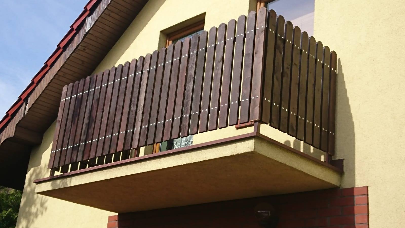 Drewniana balustrada balkonowa z pionowych desek, połączonych białymi nitami, na balkonie budynku z żółtą elewacją.