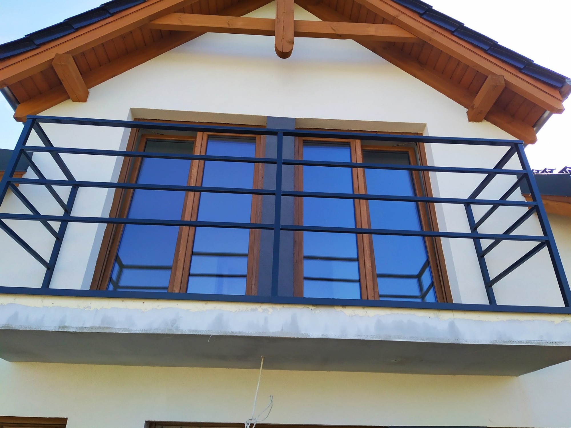 Czarna, metalowa balustrada balkonowa z prostych profili, zamontowana na balkonie z oknami w drewnianych ramach, widok od dołu na fragment elewacji domu z drewnianą podbitką dachową.