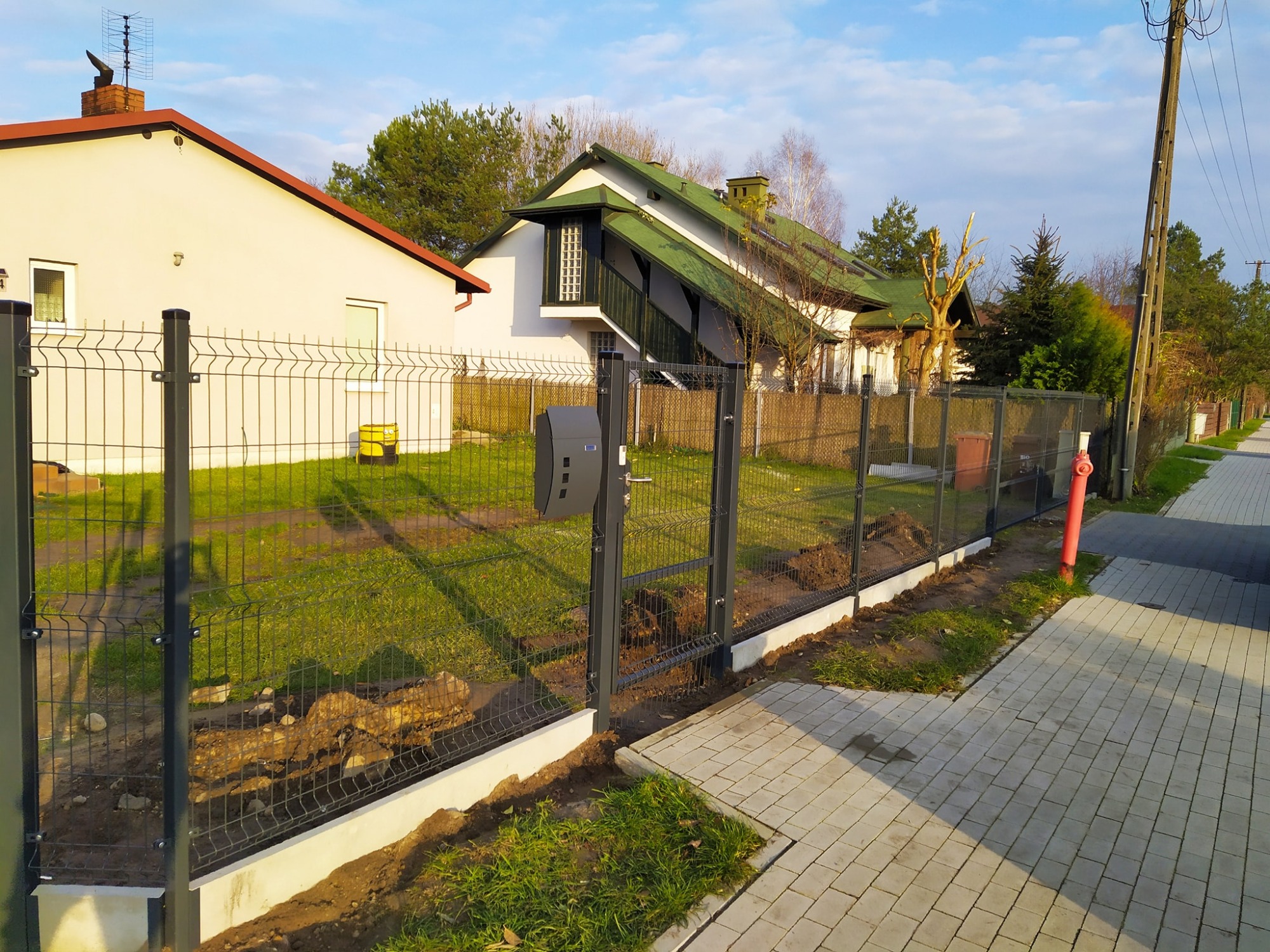 Nowo zainstalowane, grafitowe ogrodzenie panelowe z furtką i skrzynką na listy, widoczne na tle domów jednorodzinnych i chodnika z kostki brukowej w słoneczny dzień.