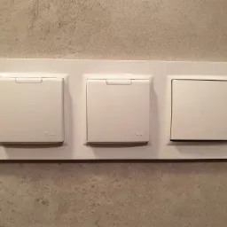 Potrójny, biały, podtynkowy włącznik z klapkami ochronnymi IP44, zamontowany na szarej, strukturalnej ścianie.
