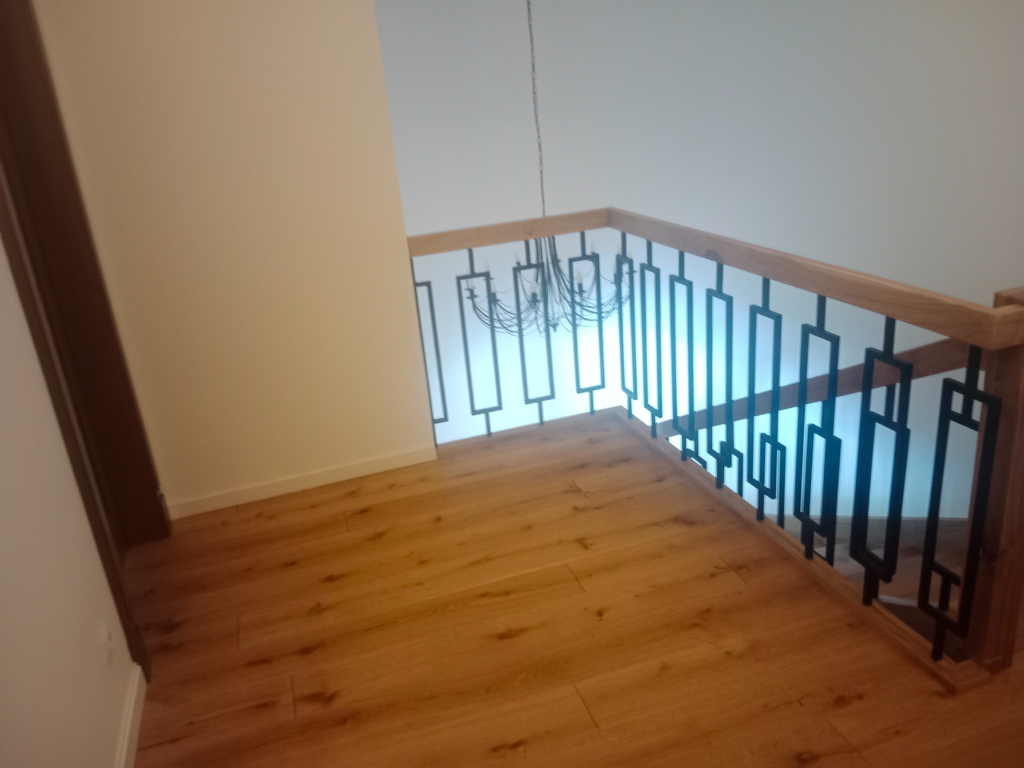 Jasne drewniane schody z czarną, geometryczną balustradą i wiszącym żyrandolem. Ujęcie z góry, podkreślające nowoczesny design i ciepłą kolorystykę drewna.