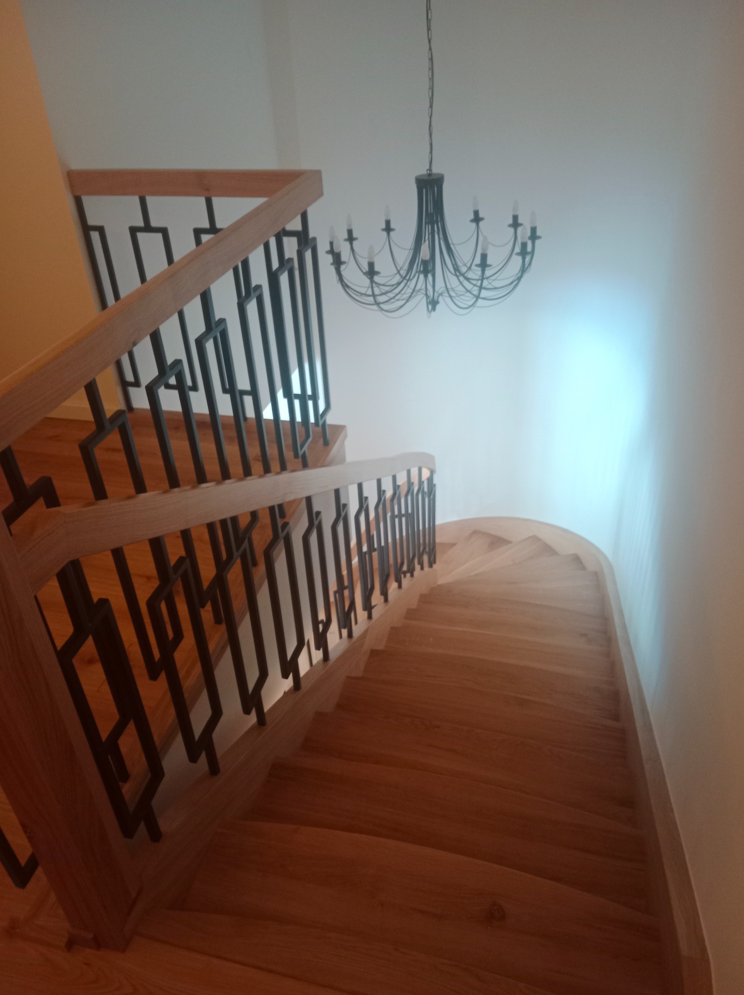Kręte schody drewniane z nowoczesną, czarną balustradą i wiszącym żyrandolem. Ujęcie z góry podkreśla zakrzywiony kształt i naturalny odcień drewna.