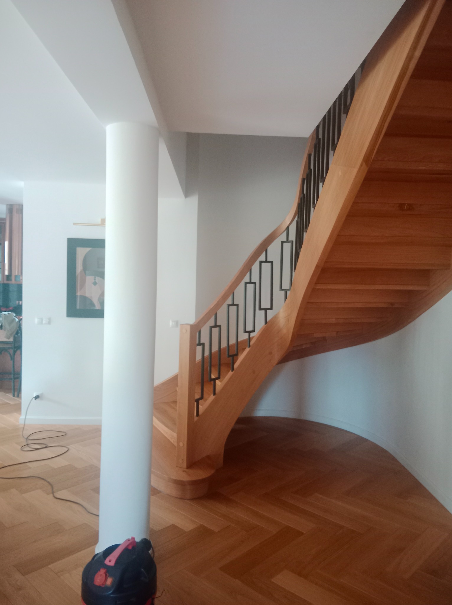 Zakręcone, jasne schody z drewnianymi stopniami i czarną, metalową balustradą, wkomponowane w jasne wnętrze z parkietem w jodełkę i kolumną.