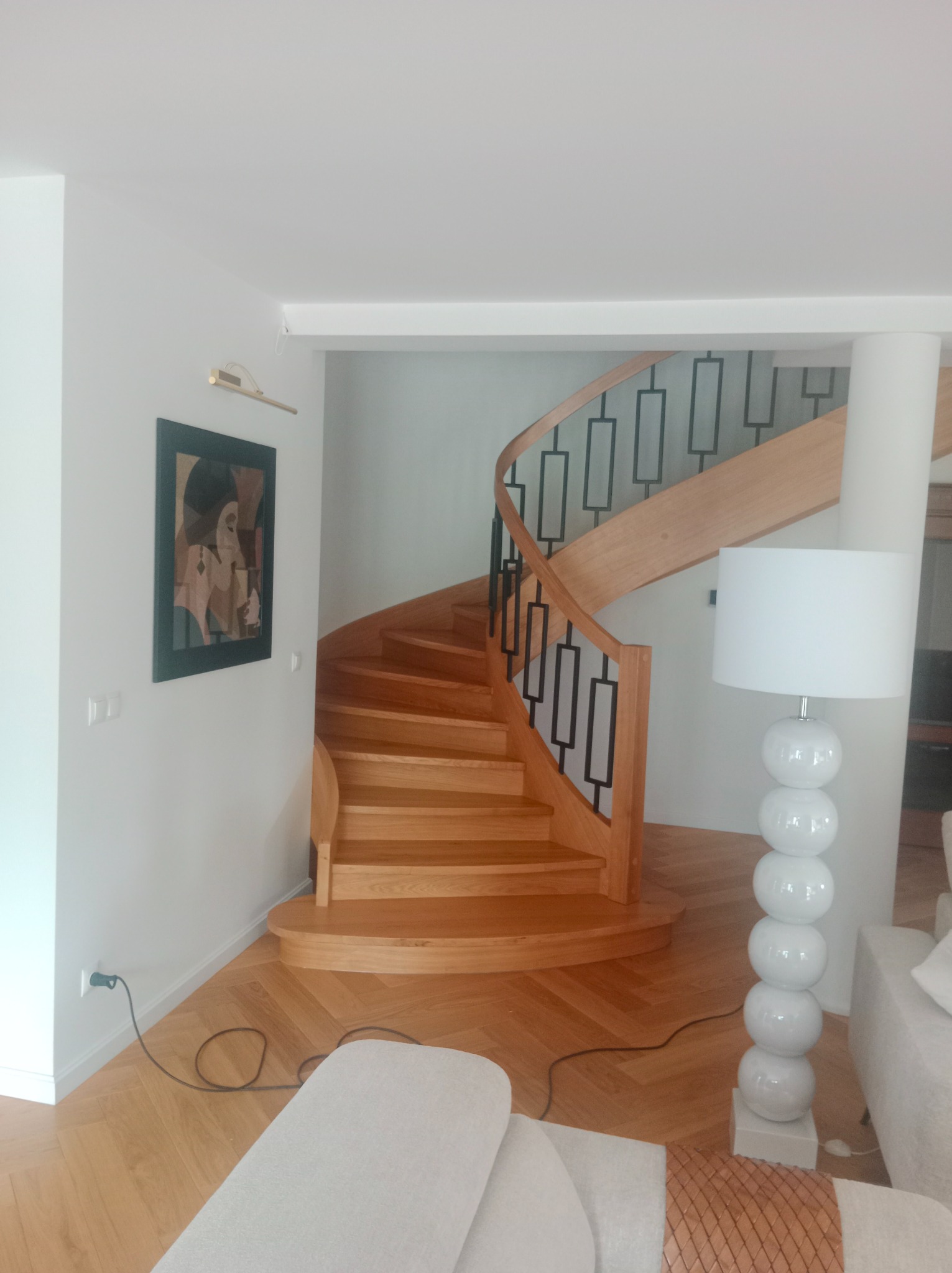 Spiralne schody z jasnego drewna z czarnymi, geometrycznymi balustradami w nowoczesnym wnętrzu z parkietem w jodełkę i lampą podłogową.