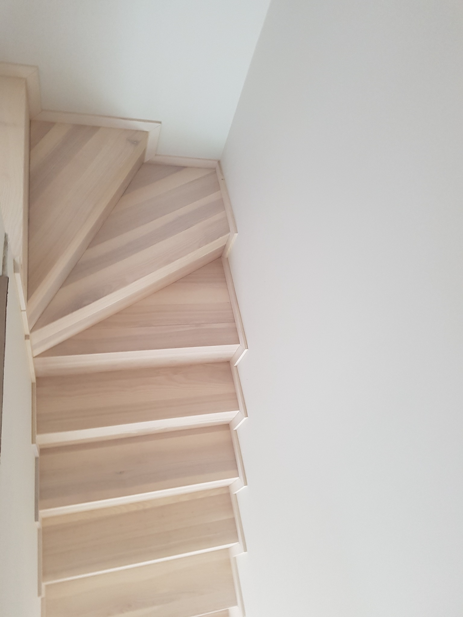 Jasne, drewniane stopnie schodów spiralnych widziane z góry, przylegające do białej ściany.