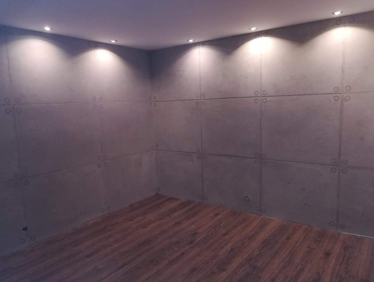 Wnętrze pokoju z panelami ściennymi imitującymi beton, połączonymi ozdobnymi śrubami, oświetlonymi punktowymi lampami sufitowymi i wykończonymi drewnopodobną podłogą.