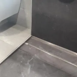 Detale wykończenia łazienki: Połączenie płytek podłogowych i ściennych z odpływem liniowym. Widoczny fragment toalety i czarna ściana.