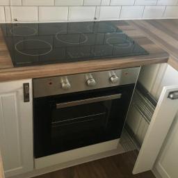 Brygada RBM - Wykończona kuchnia z białymi płytkami na ścianie, drewnianym blatem, czarną płytą indukcyjną Range Master i piekarnikiem pod spodem, widoczny fragment białej szafki z metalowym uchwytem i szklanka...