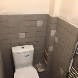 Brygada RBM - Toaleta po częściowej renowacji z szarymi płytkami imitującymi drewno, ozdobnymi wstawkami i niedokończonym fragmentem ściany.