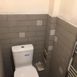 Toaleta po częściowej renowacji z szarymi płytkami imitującymi drewno, ozdobnymi wstawkami i niedokończonym fragmentem ściany.