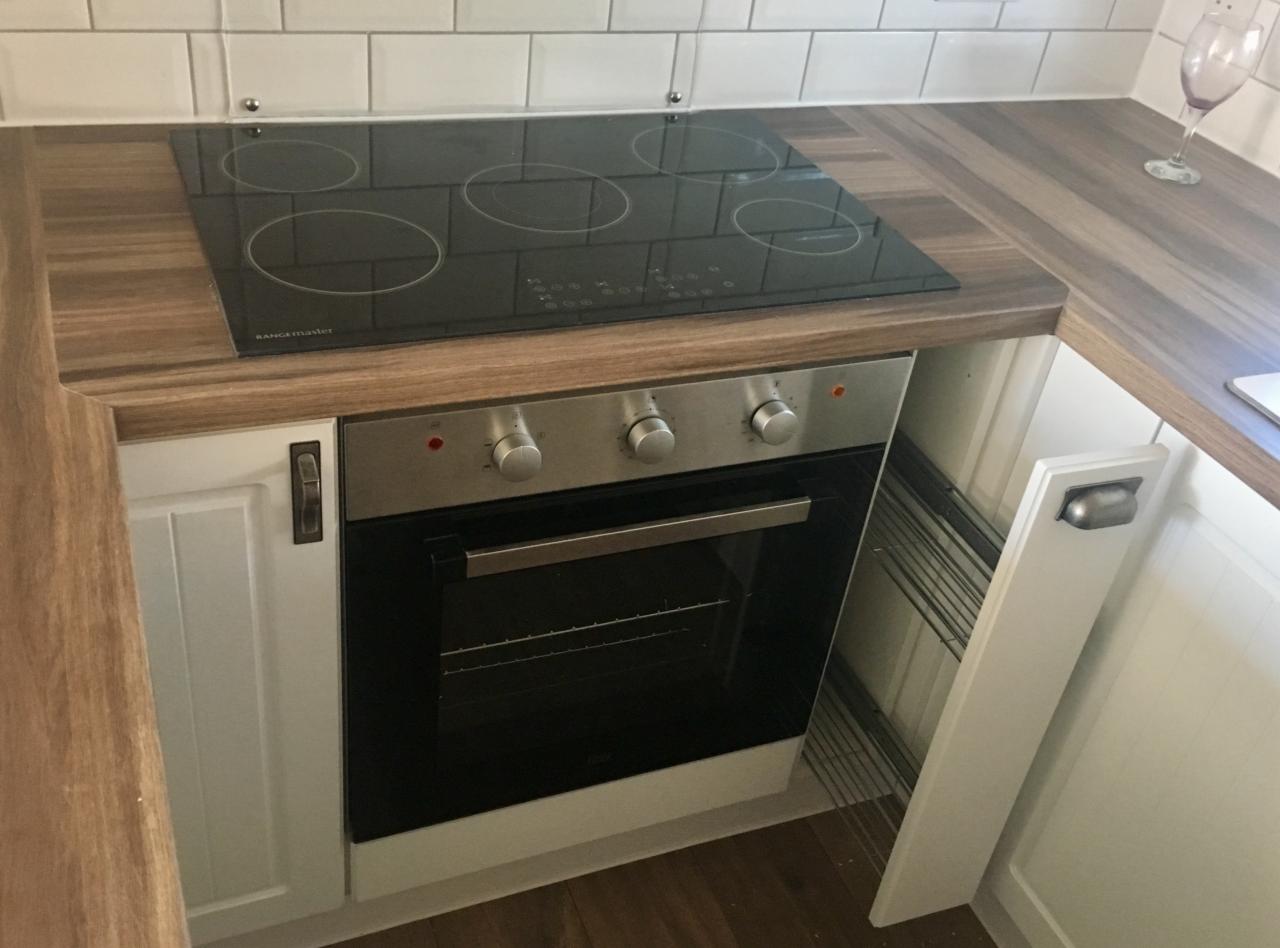 Wykończona kuchnia z białymi płytkami na ścianie, drewnianym blatem, czarną płytą indukcyjną Range Master i piekarnikiem pod spodem, widoczny fragment białej szafki z metalowym uchwytem i szklanka...