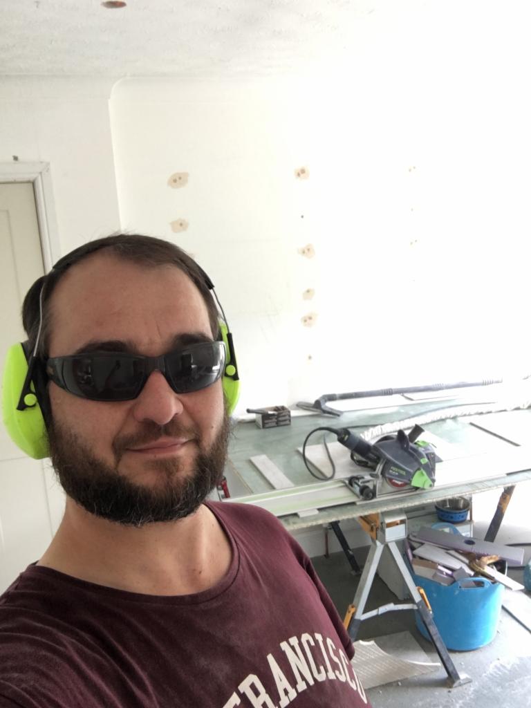 Mężczyzna w okularach ochronnych i słuchawkach, w trakcie pracy. Widoczny stół roboczy z narzędziami i materiałami. Ściana z widocznymi śladami po naprawach.