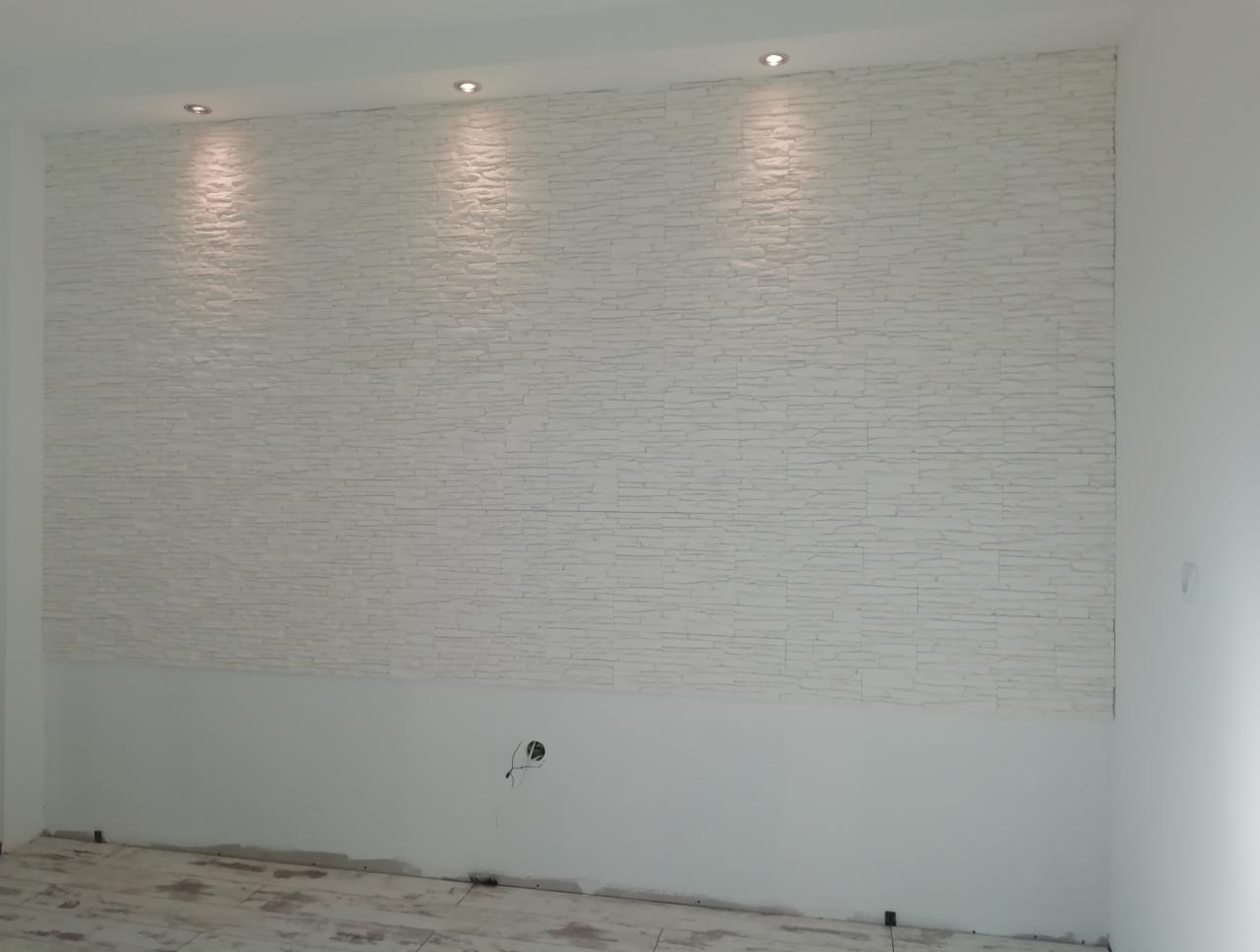 Ściana wykończona dekoracyjną, białą okładziną imitującą kamień, z widocznymi punktowymi źródłami światła w suficie i niedokończonym fragmentem ściany poniżej okładziny.