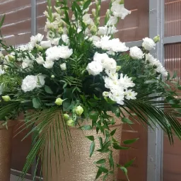 Kompozycja z białych kwiatów i zielonych liści w złotym wazonie o teksturowanej powierzchni, ustawiona przed oknem z metalową ramą.