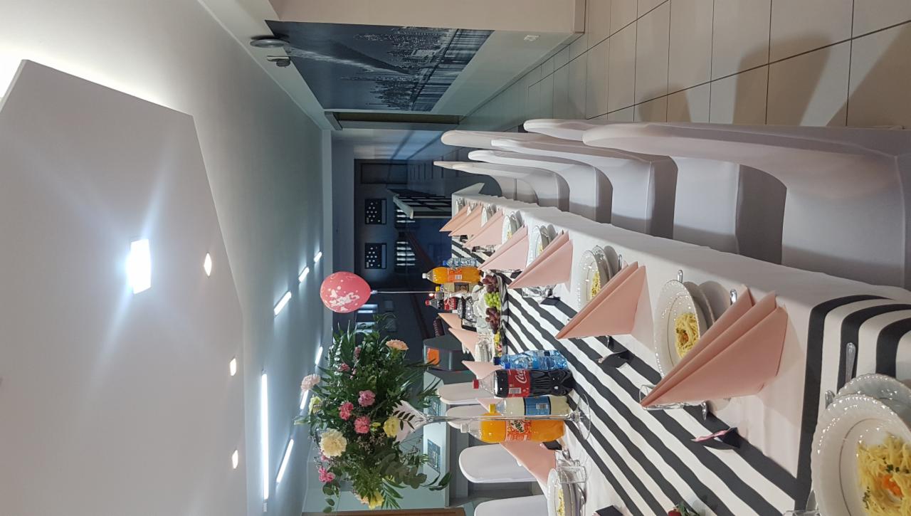 Uroczysty stół z czarno-białym bieżnikiem w paski, nakryty białymi talerzami z makaronem, różowymi serwetkami, napojami i owocami, udekorowany kwiatami i balonem z napisem, krzesła w białych...