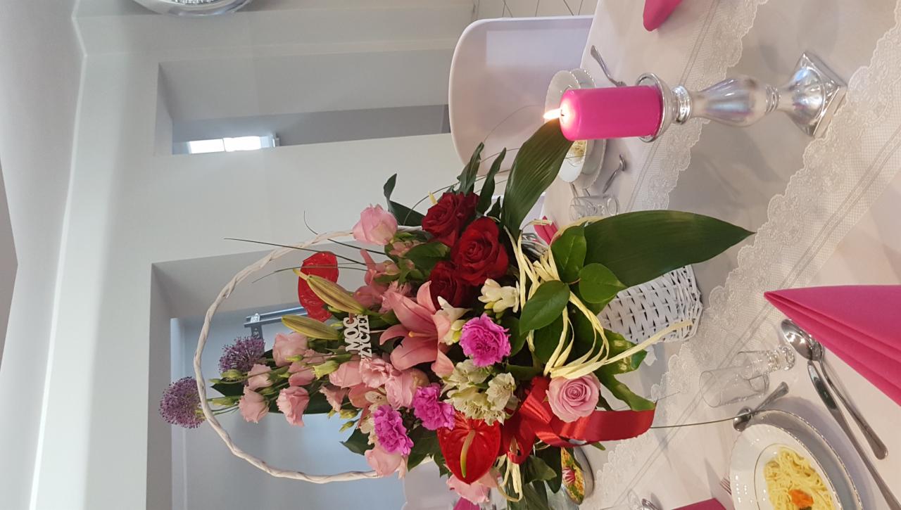 Dekoracja stołu z kwiatami w koszyku, czerwonymi anturiami, różowymi liliami i różami, świecąca różowa świeca na srebrnym świeczniku, nakrycie z koronkowym bieżnikiem i różowymi serwetkami, talerz...