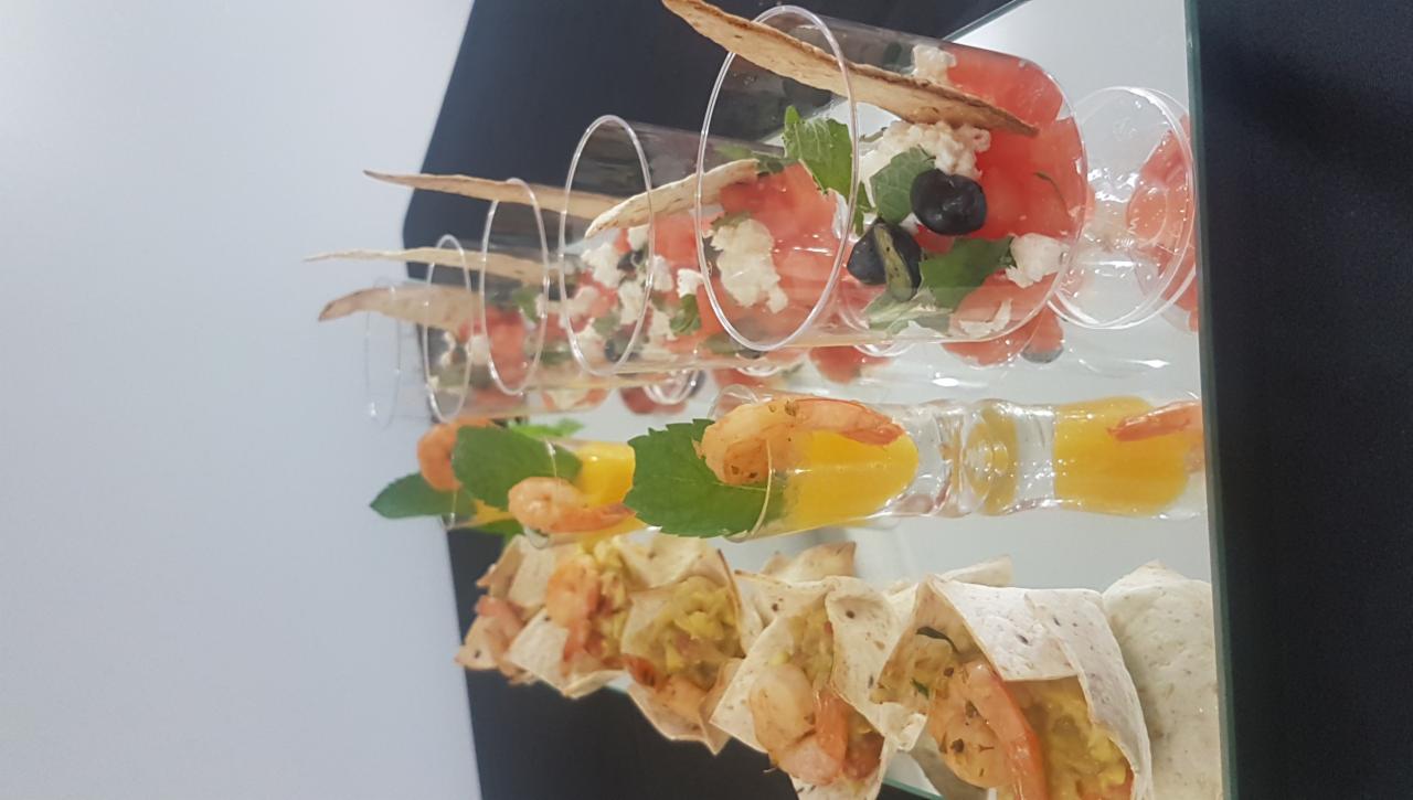 Szklane naczynia z różnorodnymi przekąskami: sałatka z fetą i arbuzem, krewetki z mango oraz mini tortille z krewetkami, ustawione rzędami na lustrzanej powierzchni.