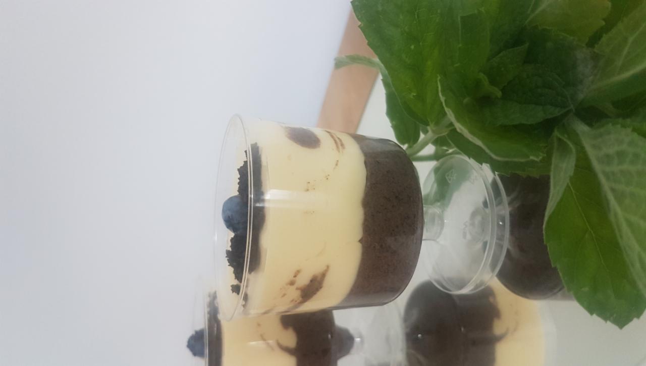 Deser warstwowy w przezroczystym pucharku, z ciemnym spodem, jasną masą i borówką na wierzchu, obok widoczne zielone liście rośliny.