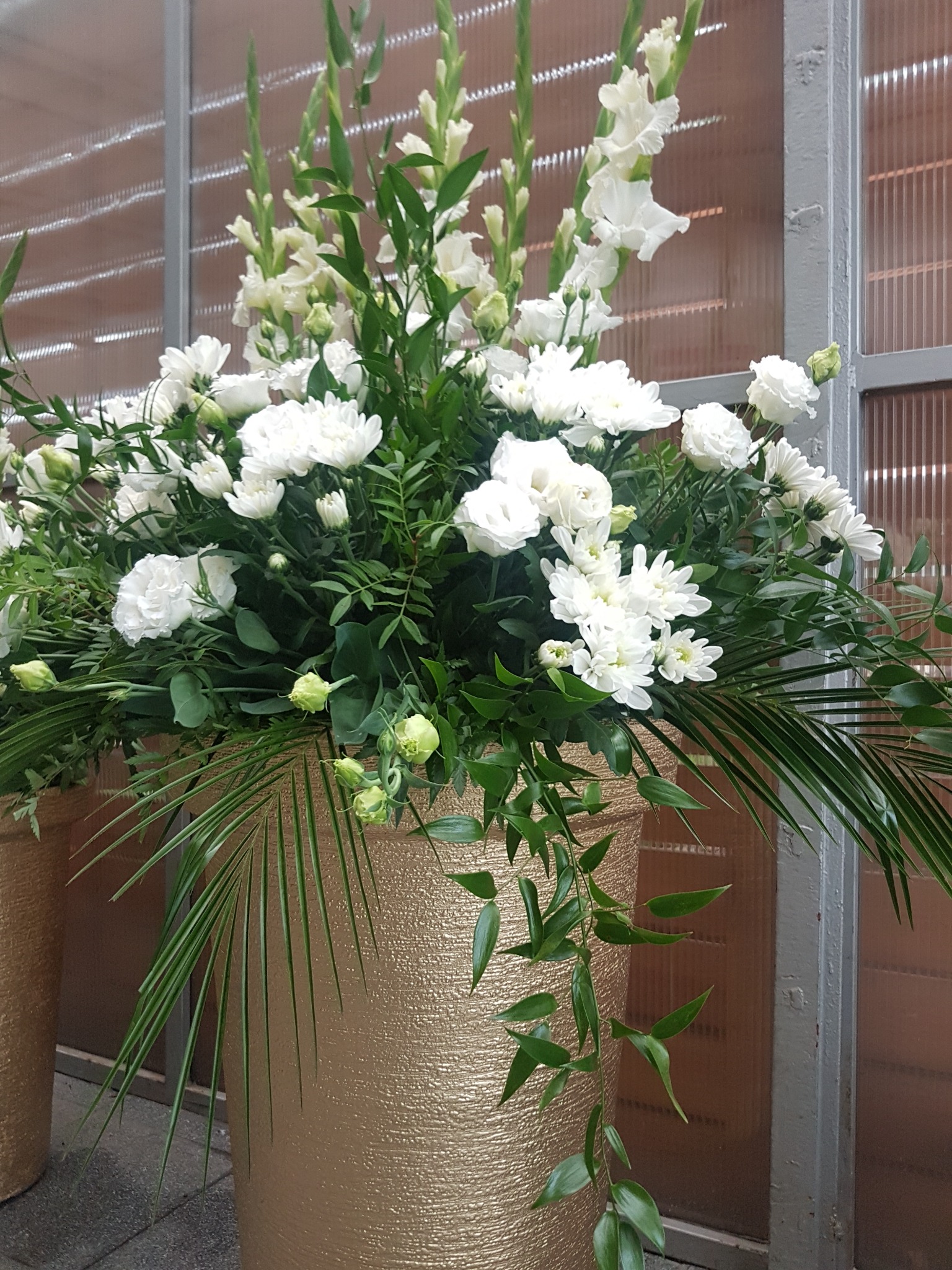 Kompozycja z białych kwiatów i zielonych liści w złotym wazonie o teksturowanej powierzchni, ustawiona przed oknem z metalową ramą.