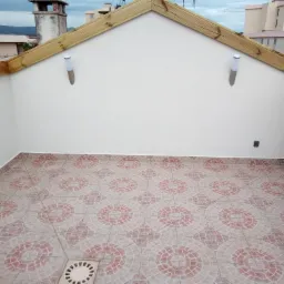 Wykończony taras na dachu z beżowo-czerwonymi płytkami w okrągłe wzory, drewnianą konstrukcją obramowania i dwiema lampami ściennymi.