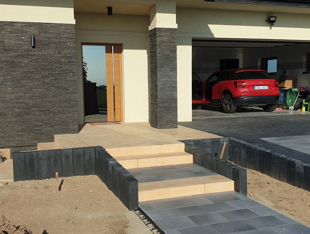 Schody wejściowe do domu z jasnych płyt, obrzeża z ciemnych elementów betonowych, w tle czerwony samochód w garażu.