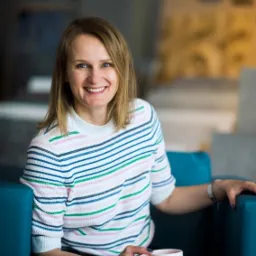 Uśmiechnięta blondynka w pasiastym swetrze, siedząca na turkusowym fotelu z kubkiem w dłoni, w tle rozmyte wnętrze biura.