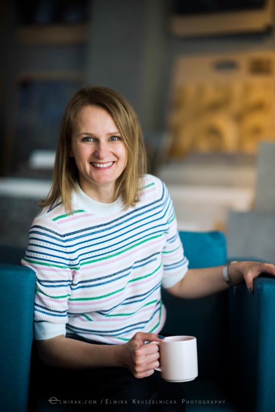 Uśmiechnięta blondynka w pasiastym swetrze, siedząca na turkusowym fotelu z kubkiem w dłoni, w tle rozmyte wnętrze biura.