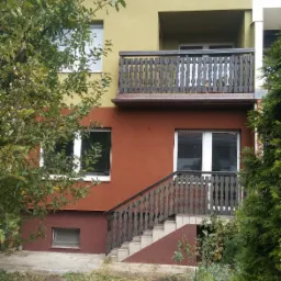 Dwukondygnacyjny dom z elewacją w odcieniach brązu i oliwki, z drewnianą balustradą balkonu i schodami wejściowymi, otoczony zielenią.