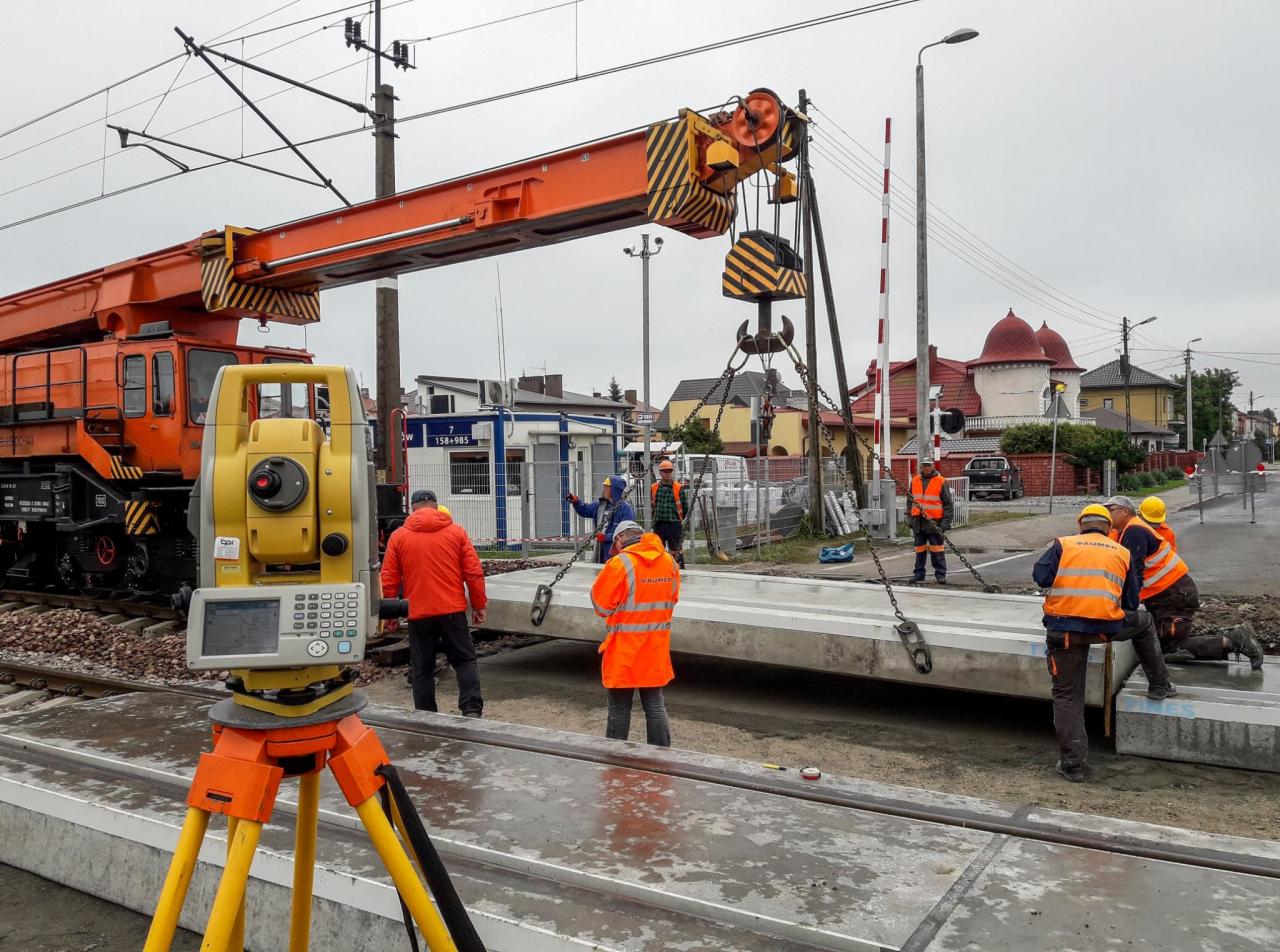 Żółty teodolit na trójnogu na pierwszym planie, w tle robotnicy w pomarańczowych kamizelkach i dźwig kolejowy podnoszący betonowy element torowiska.