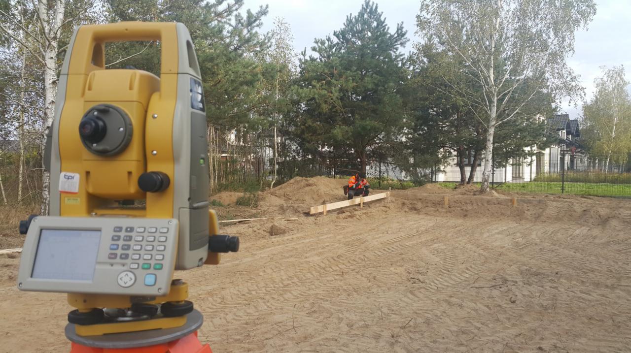 Żółty teodolit na statywie na tle placu budowy z dwoma pracownikami w pomarańczowych kamizelkach układającymi drewniane szalunki fundamentów, w tle drzewa i biały dom.