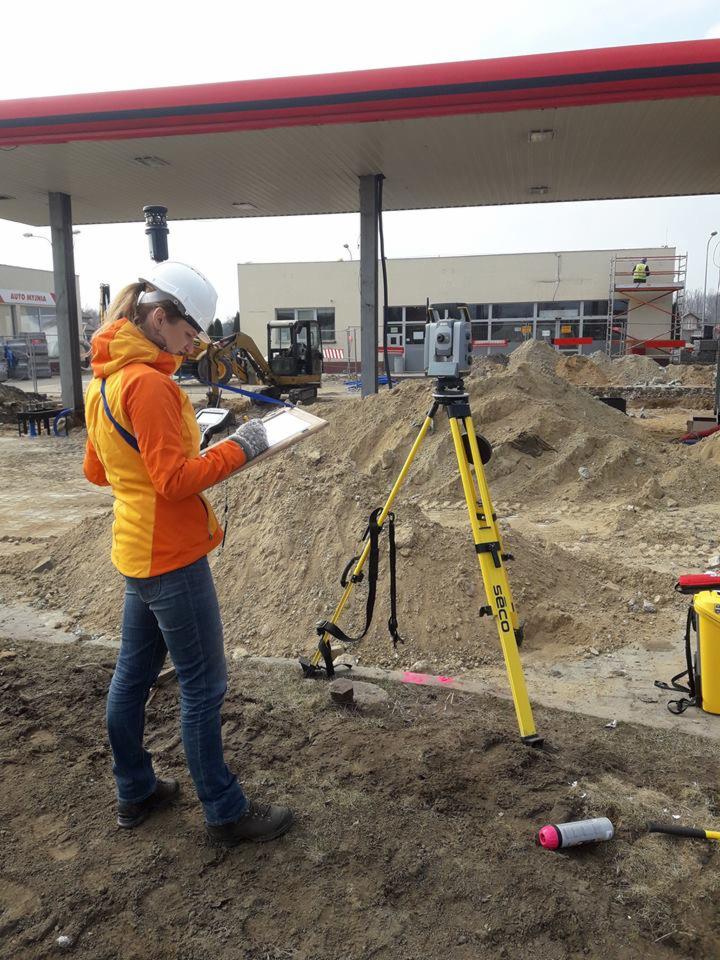 Kobieta w kasku i jaskrawej kurtce, stojąca przy niwelatorze SECO na placu budowy stacji benzynowej, sporządza notatki na tablecie.